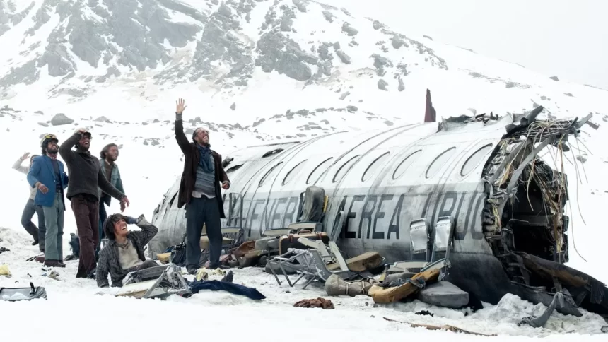 A imagem mostra parte de uma avião após queda em meio a neve com fuselagem aparente e jovens olhando para o céu pedindo socorro.