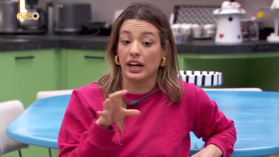Beatriz de camisa rosa, sentada na cozinha com o cabelo solto, gesticulando com a mão direita