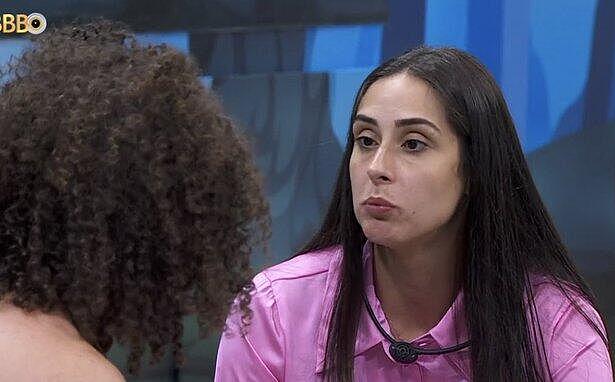 Deniziane de camisa rosa, cabelo solto, com semblante emburrado, sentada conversando com Maycon