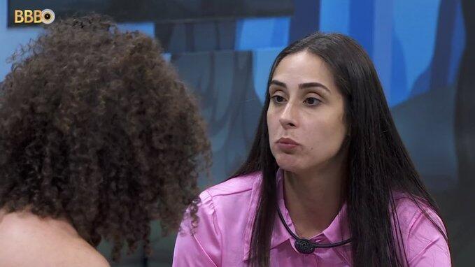 Deniziane de camisa rosa, cabelo solto, com semblante emburrado, sentada conversando com Maycon