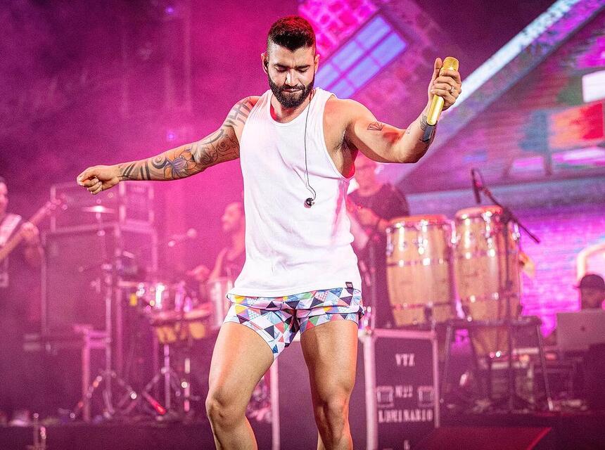 Gusttavo Lima de short curto, camisa regata branca, cantando em palco