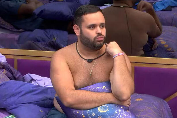 Michel sentado na cama do BBB 24, sem camisa, enrolado num lençol