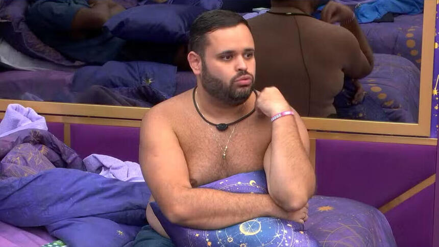 Michel sentado na cama do BBB 24, sem camisa, enrolado num lençol