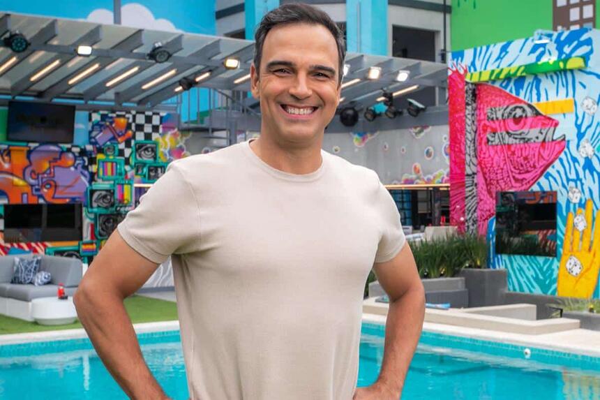 Tadeu Schmidt sorrindo, com as mãos na cintura, usando camisa cinza, em frente a piscina do BBB 24