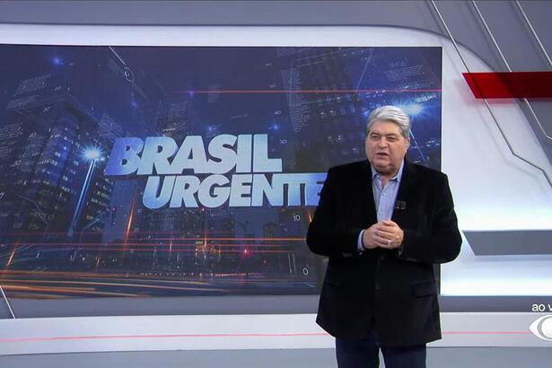 Datena apresentando o Brasil Urgente em frente ao telão com o logo do programa