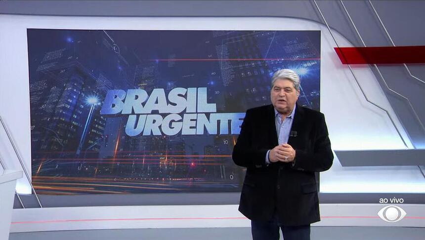 Datena apresentando o Brasil Urgente em frente ao telão com o logo do programa