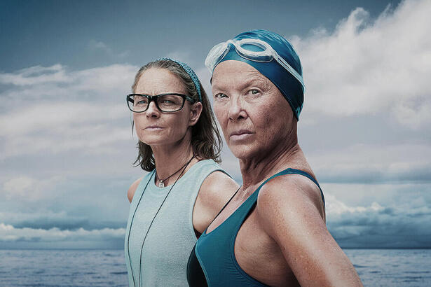 Na imagem vê-se a nadadora Diana Nyad com cerca de 60 anos com maio, touca e óculos de natação. Atrás, Bonnie, sua treinadora, aparece de blusa azul clara e óculos com o oceano de fundo.