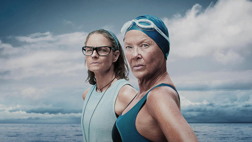 Na imagem vê-se a nadadora Diana Nyad com cerca de 60 anos com maio, touca e óculos de natação. Atrás, Bonnie, sua treinadora, aparece de blusa azul clara e óculos com o oceano de fundo.