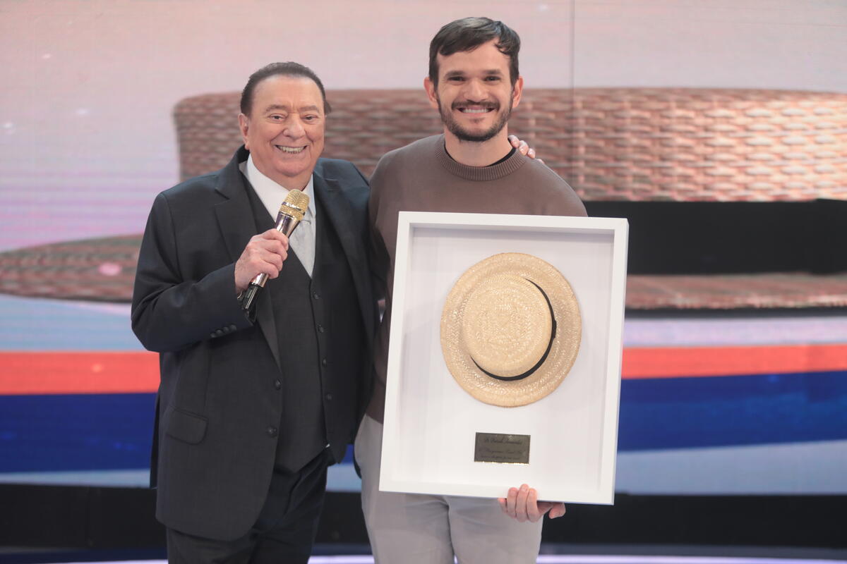 Raul Gil abraça Padre Patrick Fernandes, que segura quadro do chapéu no palco do programa do SBT 