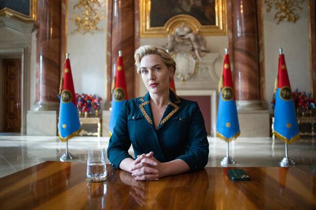 A imagem mostra Kate Winslet vestido com blaser justo azul e bordados amarelos em uma mesa oficial de um palácio europeu. Ao fundo, vê-se bandeiras ficti ícias do país da série.