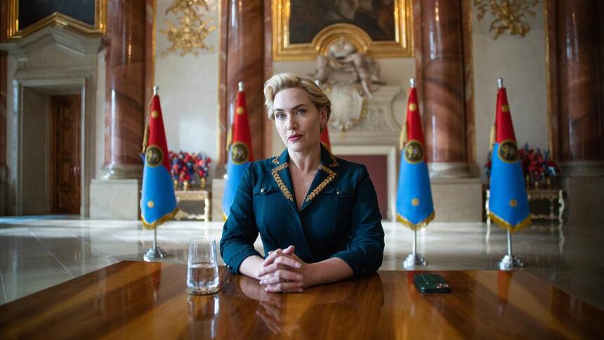 A imagem mostra Kate Winslet vestido com blaser justo azul e bordados amarelos em uma mesa oficial de um palácio europeu. Ao fundo, vê-se bandeiras ficti ícias do país da série.