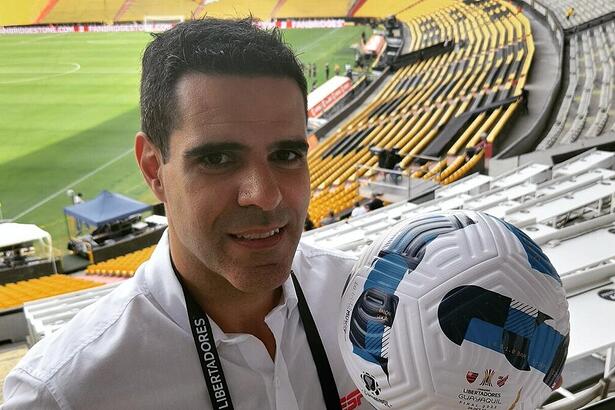 Paulo Andrade, em um estádio, nos bastidores de transmissão de um jogo na ESPN, segurando uma bola na mão