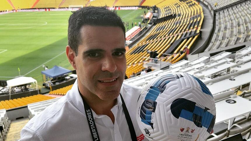 Paulo Andrade, em um estádio, nos bastidores de transmissão de um jogo na ESPN, segurando uma bola na mão