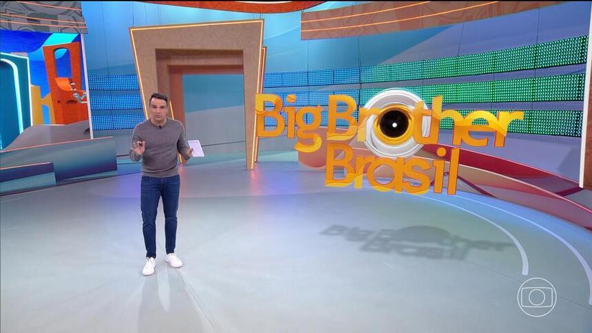 Tadeu Schmidt no centro do BBB 24, com as mãos levantadas, explicando a dinâmica da semana
