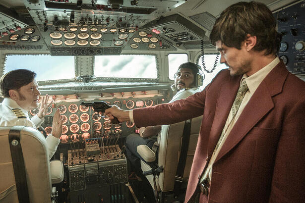 A Imagem mostra um home com terno marrom apontando uma arma para o piloto de uma avião no cockpit de uma aeronave em 1973.