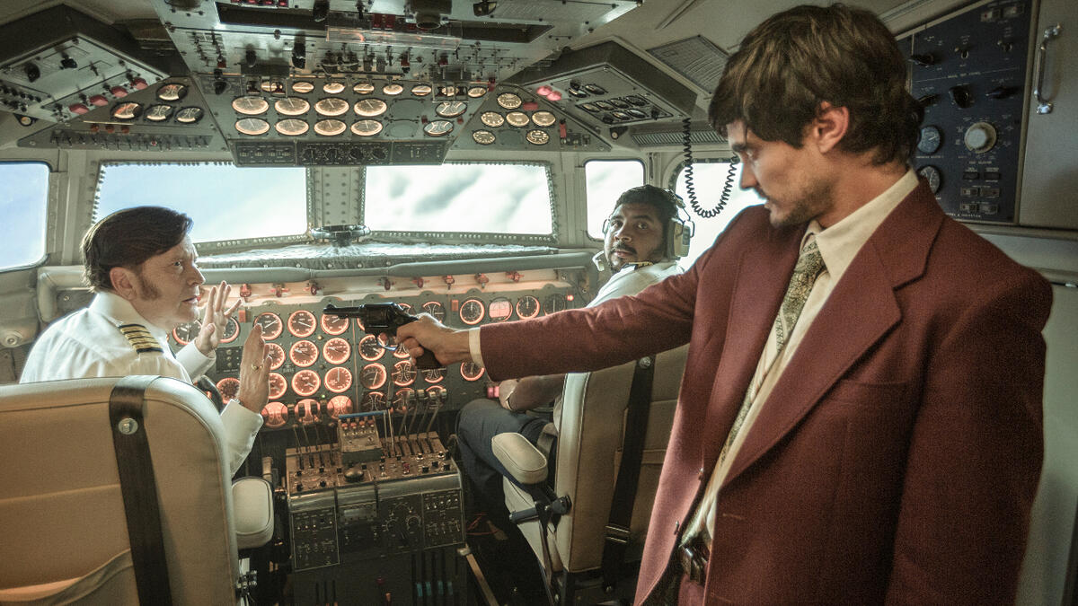 A Imagem mostra um home com terno marrom apontando uma arma para o piloto de uma avião no cockpit de uma aeronave em 1973.