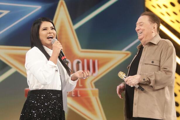 Mara Maravilha fala ao microfone sorrindo no palco do Programa Raul Gil ao lado do apresentador