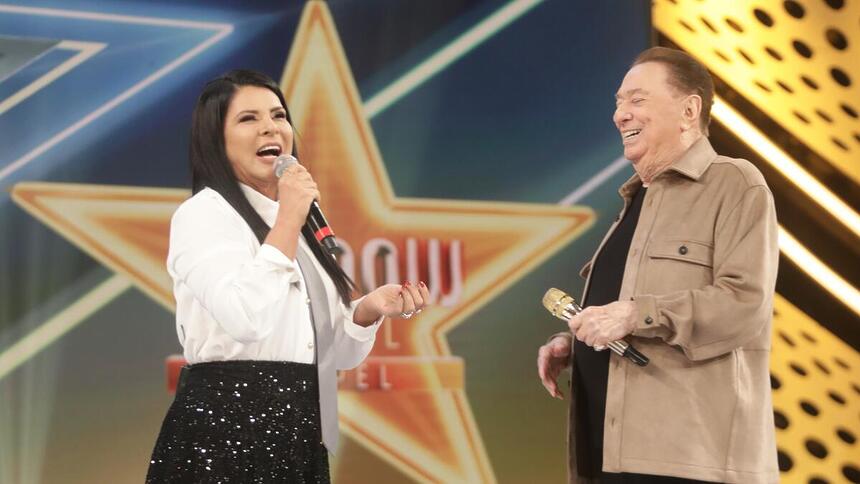 Mara Maravilha fala ao microfone sorrindo no palco do Programa Raul Gil ao lado do apresentador