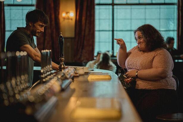 A imagem mostra um rapaz atendente de uma bar tipicamente inglês e uma mulher gorda, mais velha sentada no balcão apontando para o atendente em cena da série Bebê Rena, da Netflix