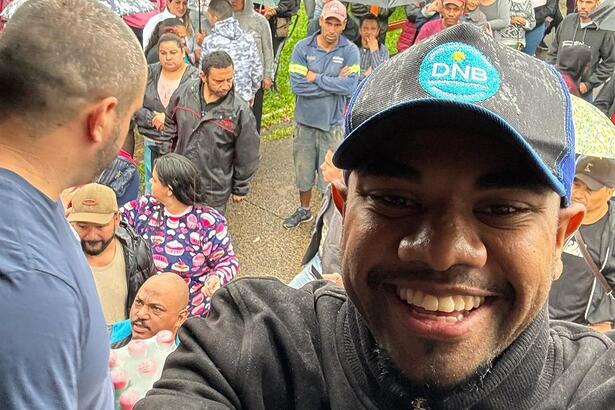 Davi sorrindo em selfie no Rio Grande do Sul, ao fundo vítimas da tragédia aparecem an foto