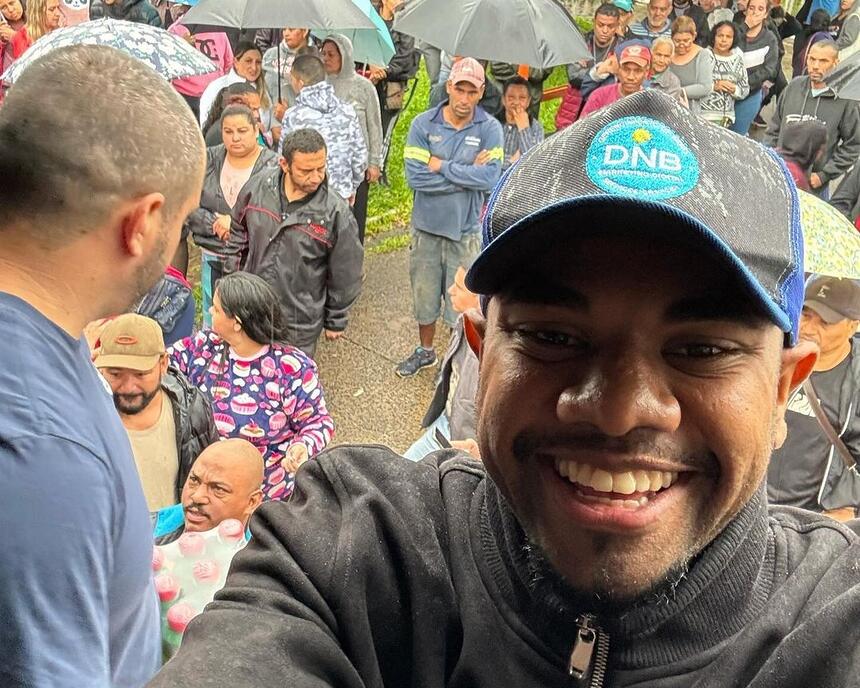 Davi sorrindo em selfie no Rio Grande do Sul, ao fundo vítimas da tragédia aparecem an foto