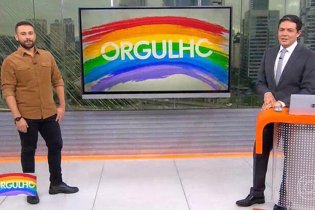 Repórter Romulo D'Ávila e o apresentador Alan Severiano em pé no cenário do SP1 em frente a uma tela de TV com o logo da série Orgulho, sobre a Parada LGBTQIA+