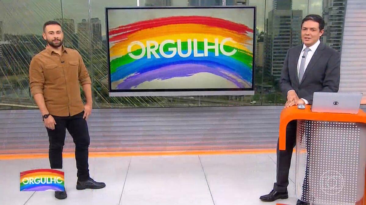 Repórter Romulo D'Ávila e o apresentador Alan Severiano em pé no cenário do SP1 em frente a uma tela de TV com o logo da série Orgulho, sobre a Parada LGBTQIA+