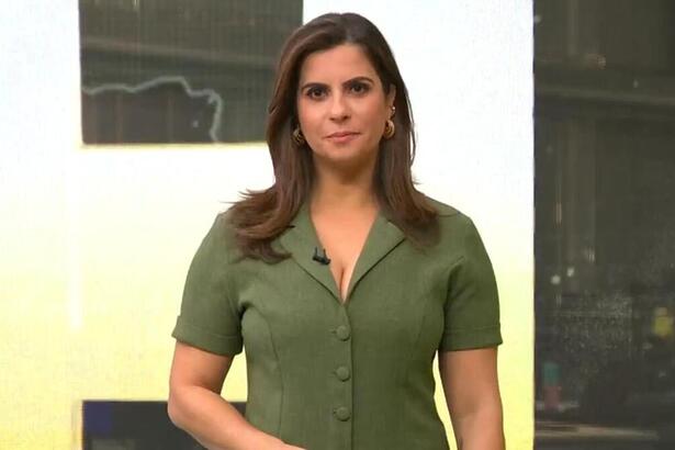 Camila Bomfim, de vestido verde, em pé em frente ao telão no estúdio do Jornal Hoje