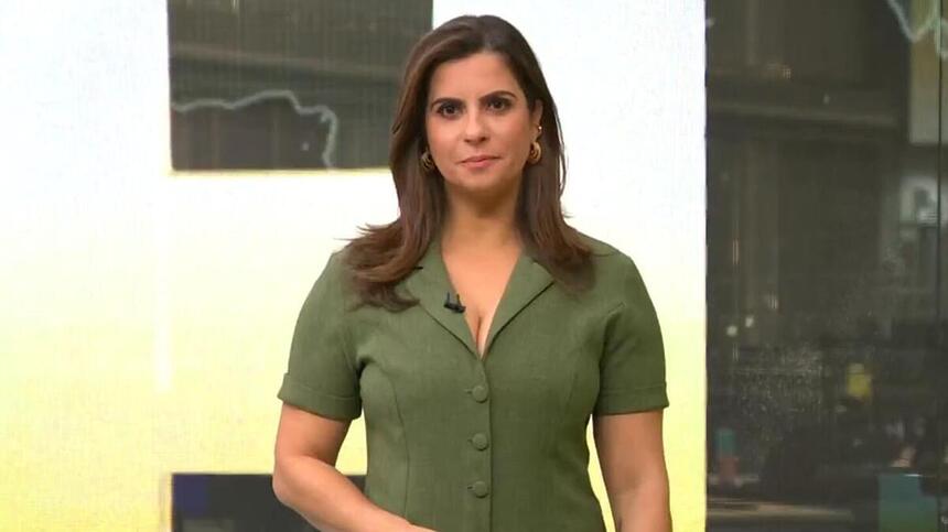 Camila Bomfim, de vestido verde, em pé em frente ao telão no estúdio do Jornal Hoje