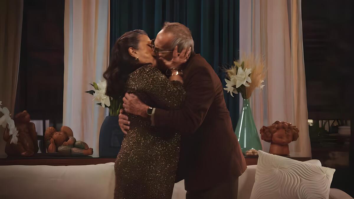 Leda (Grace Gianoukas) e Jules (Davi Pinheiro) se beijam apaixonados na sala de uma casa 