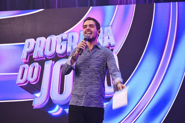 João Guilherme Silva sorrindo na frente do telão com o logo do Programa do João