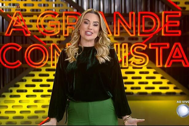 Rachel Sheherazade sorrindo na frente do logo do A Grande Conquista 2