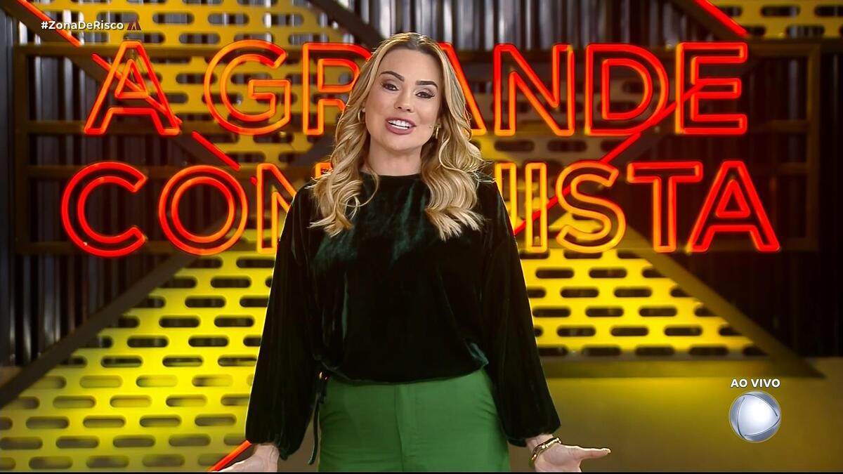 Rachel Sheherazade sorrindo na frente do logo do A Grande Conquista 2