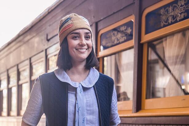 Simone Spoladore sorrindo em uma estação de trem, sorrindo em cena da novela Éramos Seis