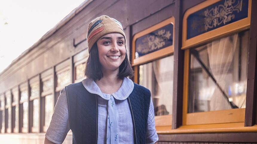 Simone Spoladore sorrindo em uma estação de trem, sorrindo em cena da novela Éramos Seis