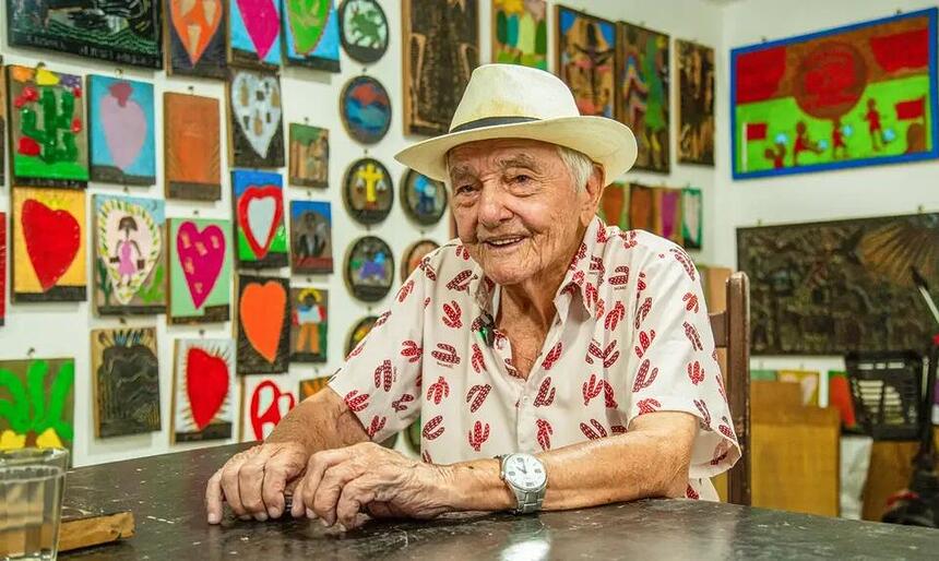 Xilogravurista pernambucano J. Borges sentado em uma mesa com chapéu branco na cabeça