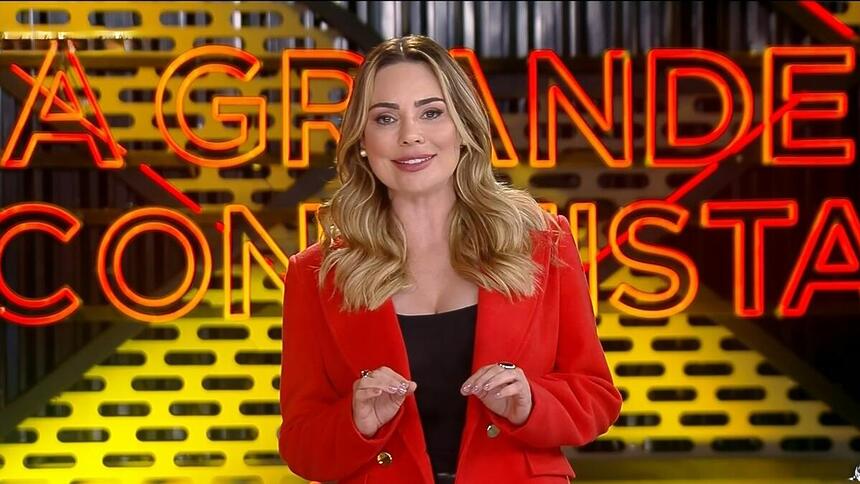 Rachel Sheherazade veste um casaco vermelho e sorrir enquanto apresenta o A Grande Conquista 2