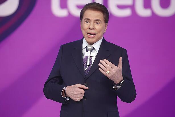 Silvio Santos com o braço esquerdo levantado no cenário do SBT