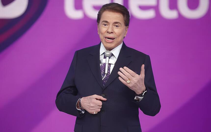 Silvio Santos com o braço esquerdo levantado no cenário do SBT