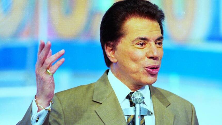 Silvio Santos levanta o braço esquerdo e carrega um microfone no peito