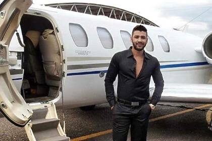 Gusttavo Lima em frente a um avião de pequeno porte. Ele veste camisa preta e está sorridente