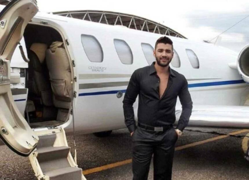 Gusttavo Lima em frente a um avião de pequeno porte. Ele veste camisa preta e está sorridente