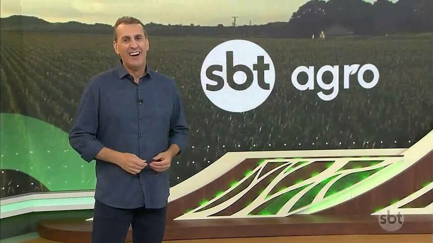 Apresentador Sandro Ivanowski no cenário do SBT Agro, em frente ao telão e sorrindo