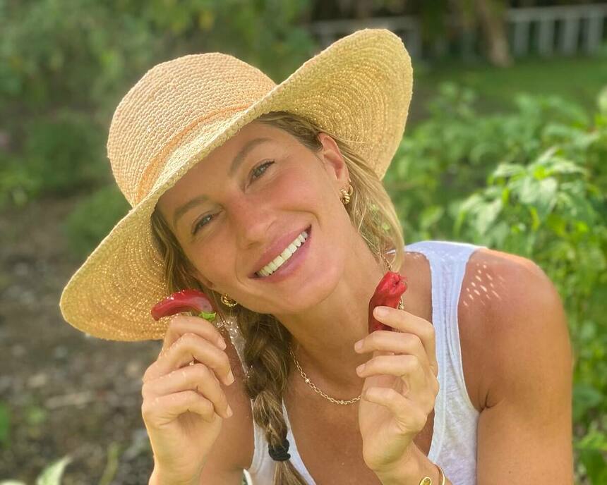 Gisele Bündchen segura duas frutas nas mãos e sorrir com chapéu de camponesa