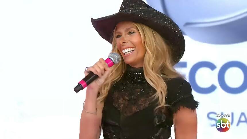 Adriane Galisteu sorrindo no palco do Teleton com o logo da RECORD ao fundo