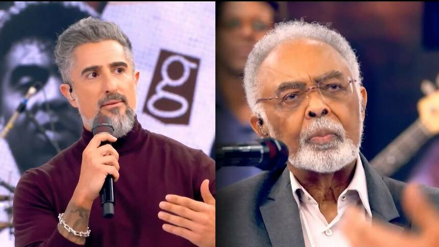 Marcos Mion de blusa vermelha falando com Gilberto Gil no palco do Caldeirão, que está com um olhar sério