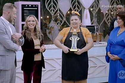 Eli, com o avental preto e o troféu, ao lado dos jurados Andre Mifano e Caroline Crema e a apresentadora Fabiana Karla no Bake Off Brasil