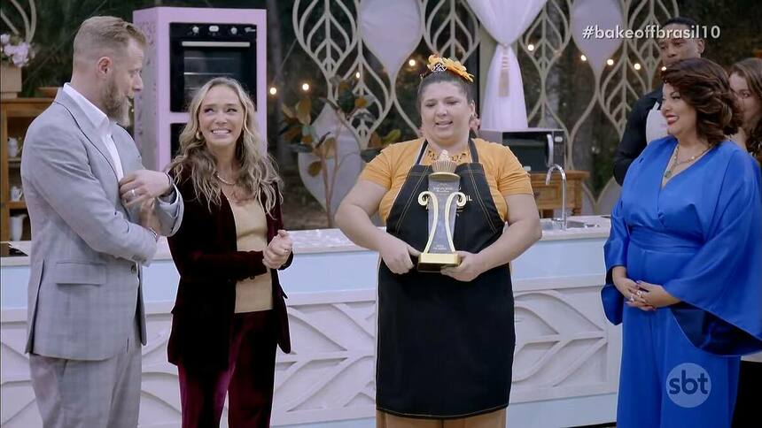 Eli, com o avental preto e o troféu, ao lado dos jurados Andre Mifano e Caroline Crema e a apresentadora Fabiana Karla no Bake Off Brasil