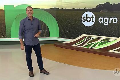 Sandro Ivanowski sorridente e em pé no centro do cenário do SBT Agro, ele vest calça preta e camisa azul