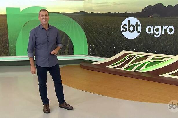 Sandro Ivanowski sorridente e em pé no centro do cenário do SBT Agro, ele vest calça preta e camisa azul